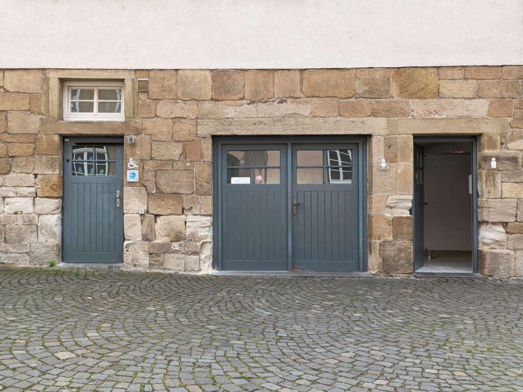 Links die Tür zur barrierefreien Toilette, in der Mitte der Eingang zur Damentoilette, rechts der Eingang zur Herrentoilette.