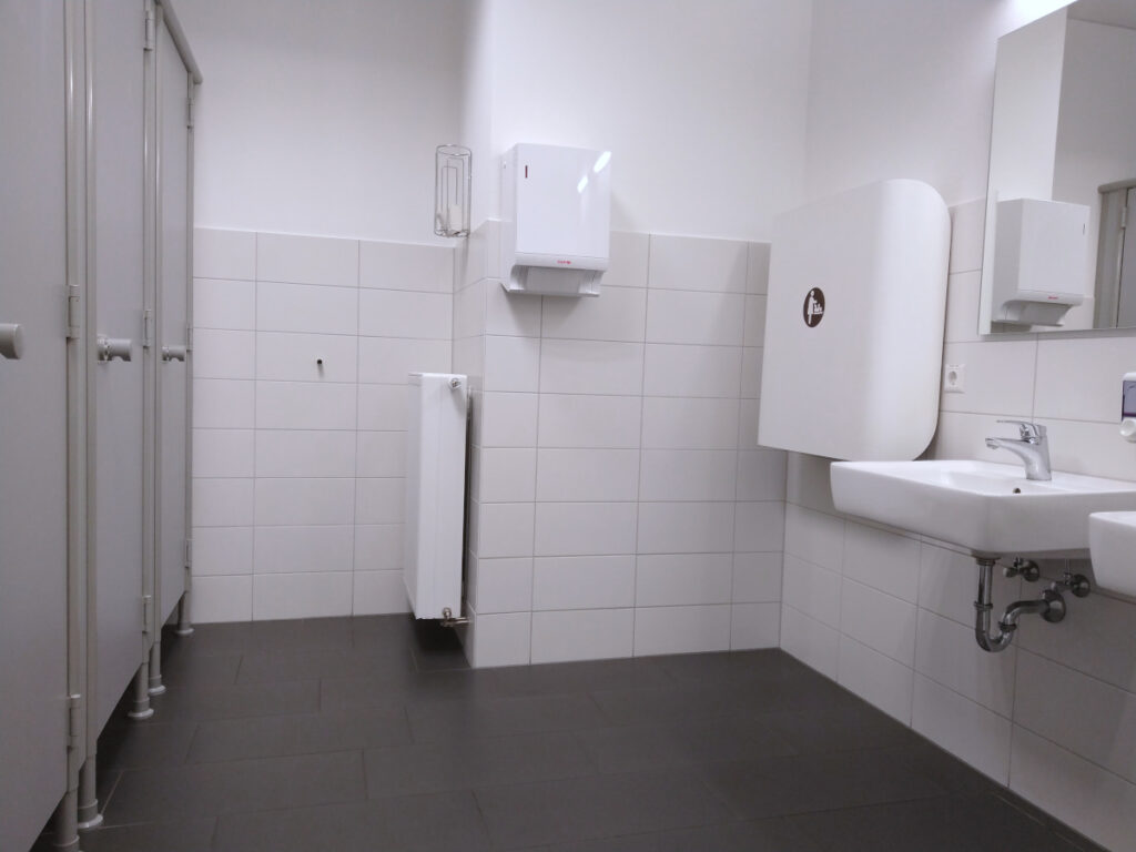 Toilette mit weißen Wandfliesen und dunklen Bodenfliesen. Links drei Toilettenkabinen, rechts Waschbecken und an der rechten Wand ein klappbarer Wickeltisch.