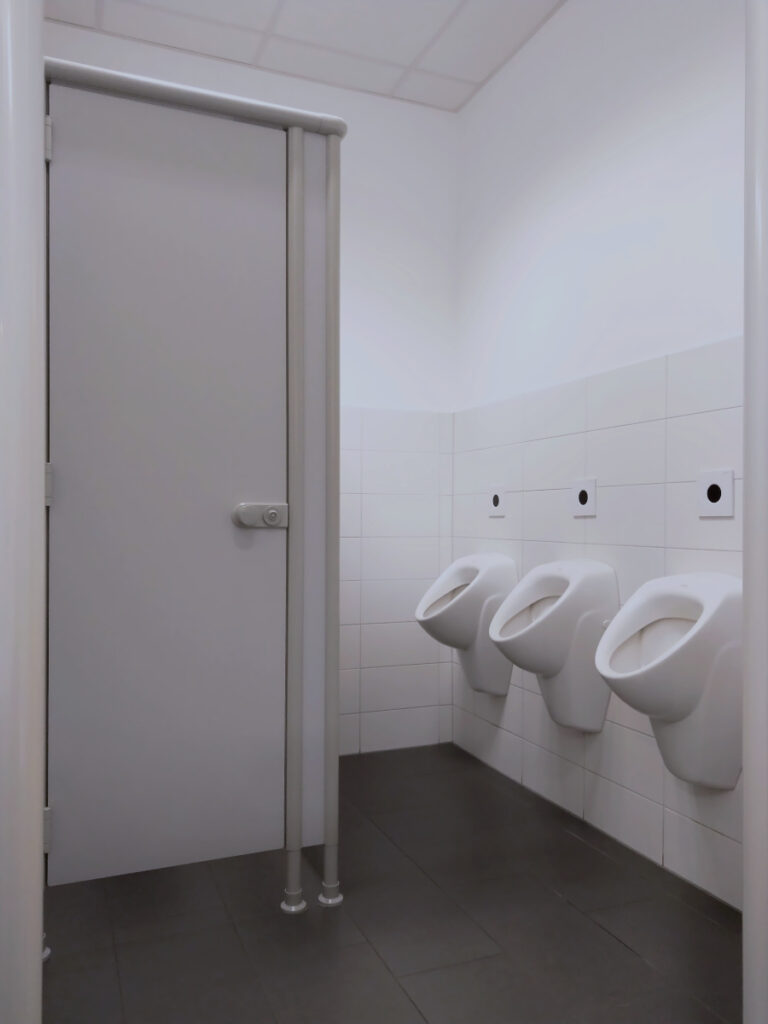 Toilette mit weißen Wandfliesen und dunklen Bodenfliesen. Geradeaus eine Toilettenkabine, rechts drei Pissoirs.