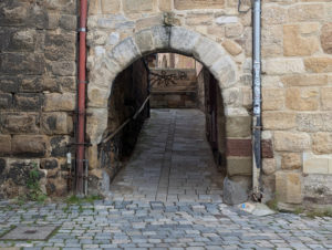 Steinerner Torbogen, der in eine gepflasterte, enge Gasse führt, die zwischen alten Sandsteinmauern verläuft.