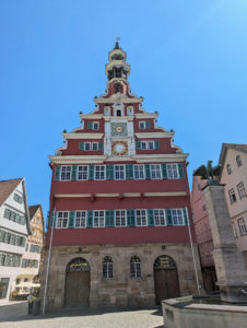 Historisches Rathaus mit rot-weißer Fassade und verziertem Turm, auf dem sich eine Uhr befindet. Im Vordergrund ein Brunnen mit Adlerfigur.