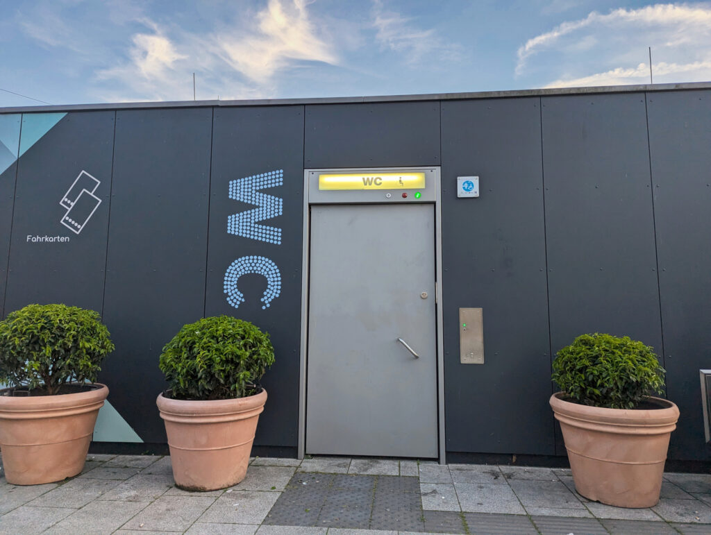 Einstöckiger Container mit Tür, darüber ein Feld mit der Aufschrift „WC“. Rechts vom Türgriff befindet sich ein Schlüsselloch für das automatische Öffnen der Tür.