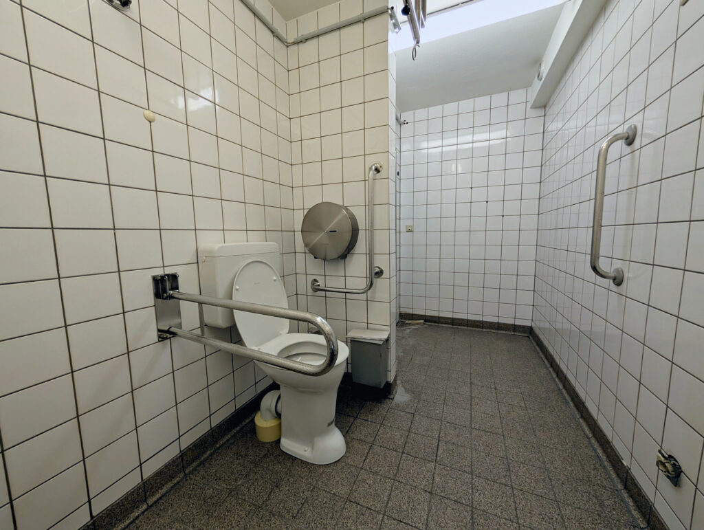 WC mit Standfuß, Klappgriff links und fester Griff an der Wand rechts. Spülkasten hinten.