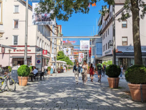 Fußgängerzone mit vielen bummelnden Menschen. Am Rand Bäume, die Schatten spenden, und bunte Plakate, die hoch oben, quer über die Straße aufgehängt sind.