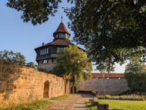 Zentraler runder Turm mit steinernem Sockel und hölzernem Aufbau, von mittelalterlichen Mauern flankiert; ein Weg führt direkt zum Turmeingang.