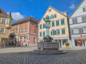 Im Vordergrund ein runder Brunnen. Auf dem Brunnenrand ein Relief mit Menschen in einem Wirtshaus. In der Mitte des Brunnen ragt eine Säule empor auf der ein Reiter steht, der in ein Horn bläst. Im Hintergrund mehrere Häuser. Platz mit Kopfsteinpflaster.