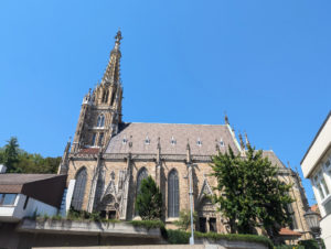 Außenansicht einer gotischen Kirche mit einem reich verzierten Kirchturm. Die Fassade weist zahlreiche dekorative Elemente auf, darunter detaillierte Reliefs über zwei seitlichen Toren.