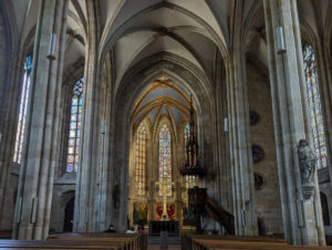 Innenansicht einer gotischen Kirche mit hohen Spitzbogenfenstern, leuchtenden Buntglasfenstern im Altarbereich, schlanken Säulen, und verzierter Holzkanzel.