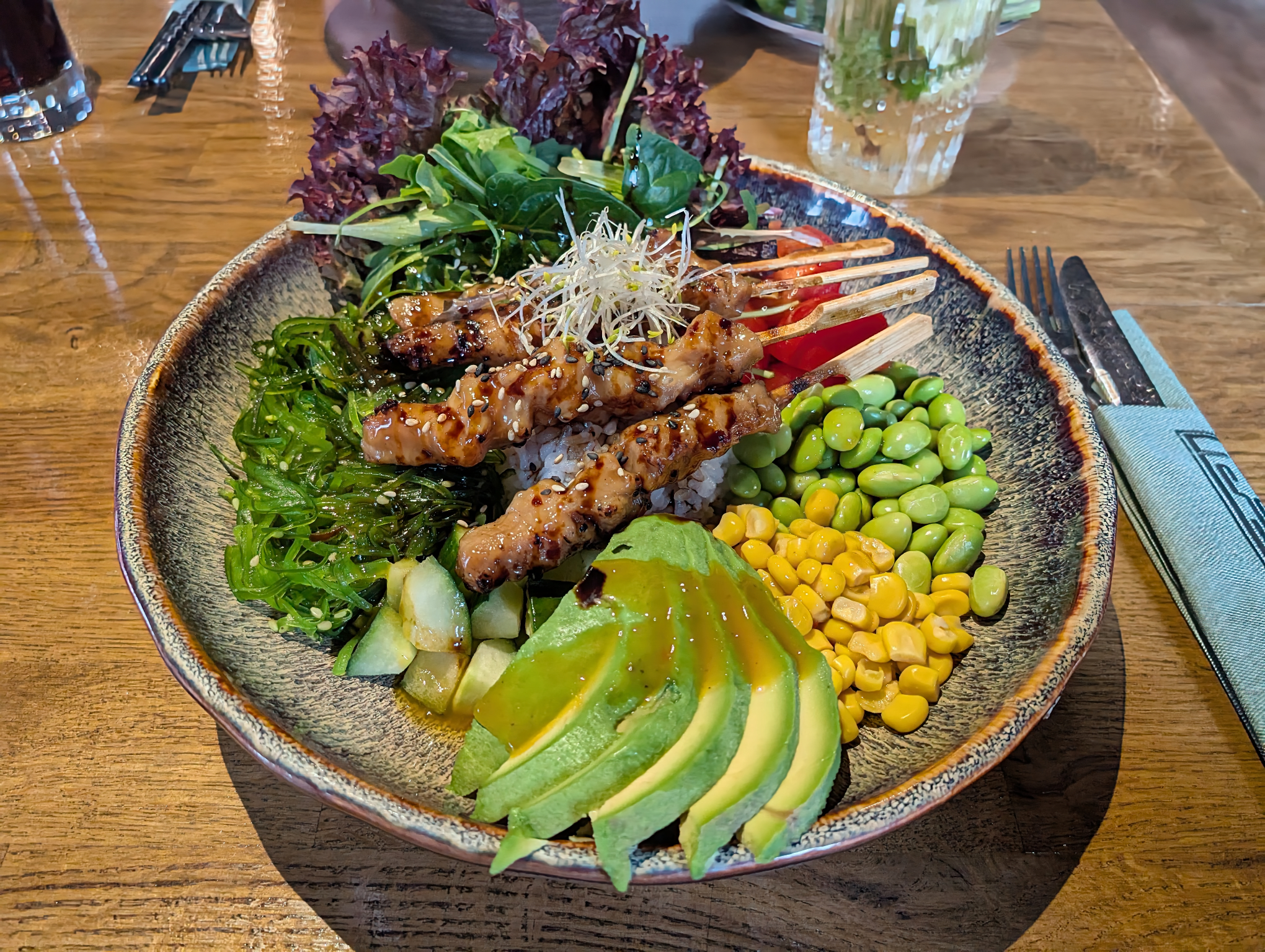 Schale mit Avocado, Mais, Edamame, Reis und Blattsalat, darüber vier gegrillte Fleischspieße quer gelegt.