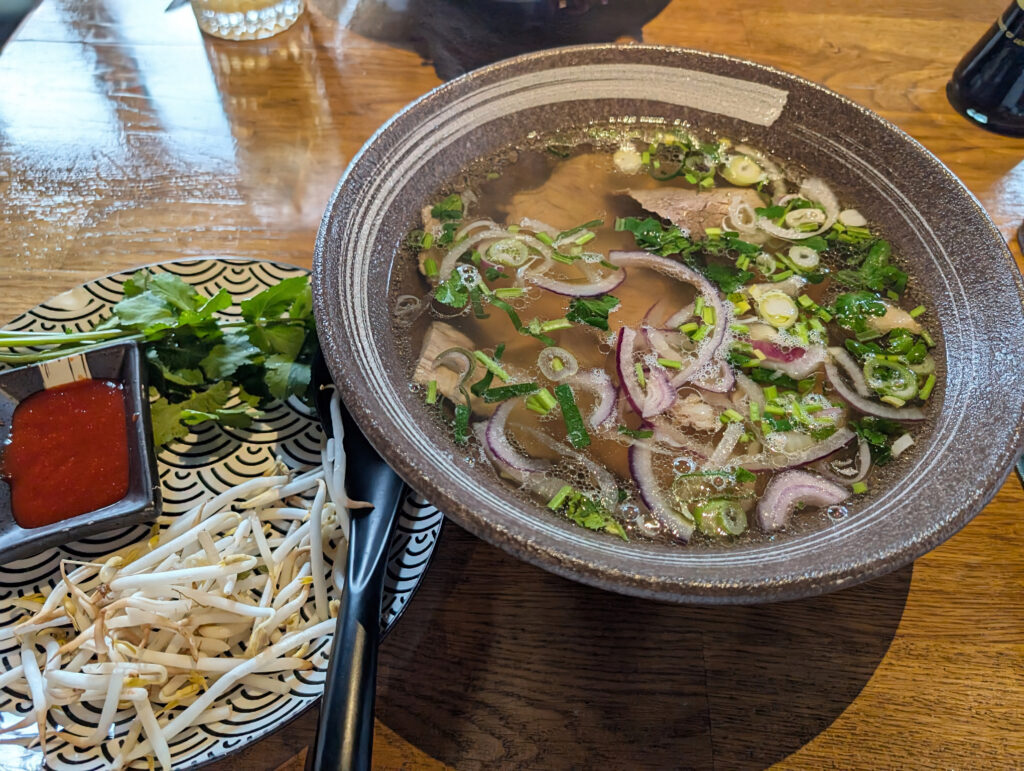 Rechts eine Schale mit Suppe, auf der Oberfläche schwimmen rote Zwiebeln, Petersilie und Frühlingszwiebeln. Daneben ein Teller mit einer Schale roter Soße, Sprossen und Koriander.