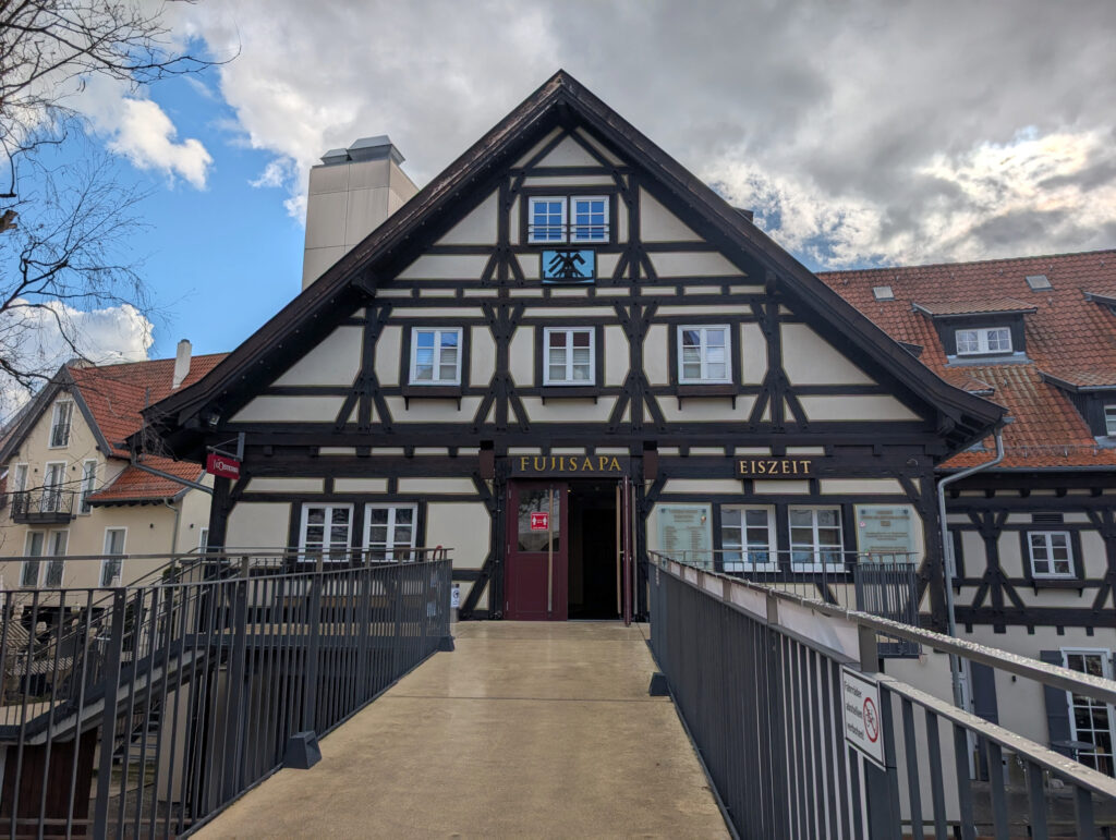 Steg mit Metallgeländer führt zu einer geöffneten Tür in der oberen Etage eines Fachwerkhauses.