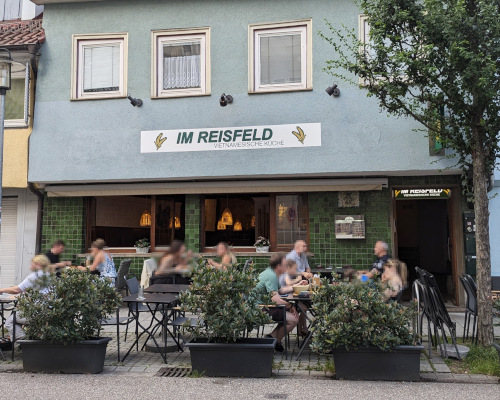 Außen-Ansicht vom Restaurant "Im Reisfeld". Außen-Wand mit grünen Fliesen. 2. Stock blau gestrichen. Vor dem Gebäude, neben dem Gehweg: mehrere Tische und Stühle, auf denen Gäste sitzen. Als Abgrenzung zur Straße mehrere Pflanzen in Kübeln.