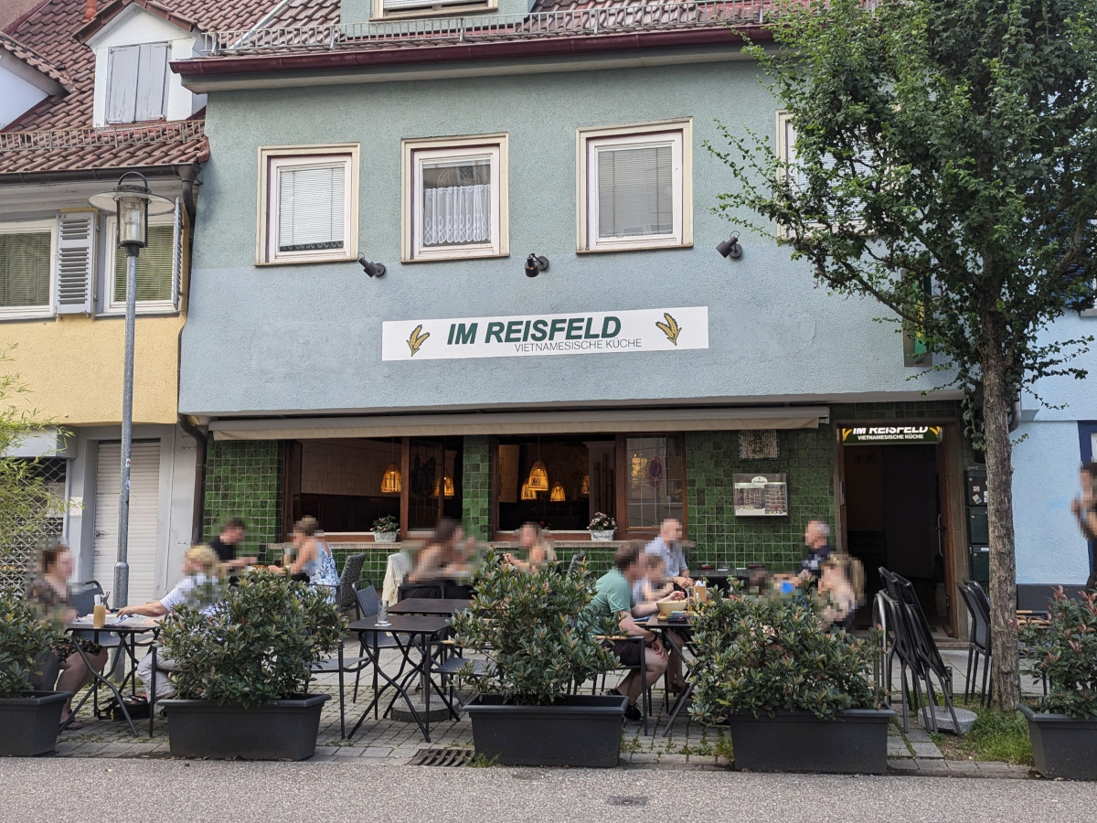 Außen-Ansicht vom Restaurant "Im Reisfeld". Außen-Wand mit grünen Fliesen. 2. Stock blau gestrichen. Vor dem Gebäude, neben dem Gehweg: mehrere Tische und Stühle, auf denen Gäste sitzen. Als Abgrenzung zur Straße mehrere Pflanzen in Kübeln.