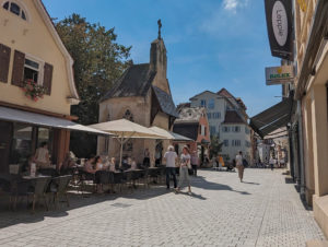 Einkaufsstraße auf einer Brücke. Am linken Brückenrand kleine Häuschen mit einem Café, einer Kapelle und einem Laden. Mehrere Menschen, die in Sommerkleidung entlang flanieren und einige die im Café sitzen.