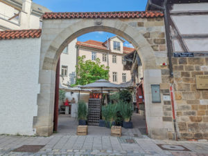 Malerischer Innenhof-Eingang mit rundbogigem Tor, das zu einer Sektbar im Freien mit Sitzplätzen, Pflanzen und Sonnenschirmen führt.