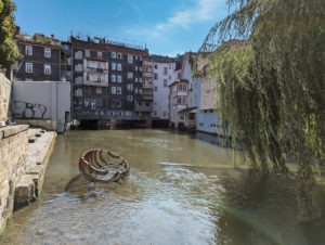 Breiter Wasserkanal umrahmt von Gebäuden. Darunter eine Häuserreihe quer über den Kanal. Mitten im Kanal eine Skulptur in Form eines Schiffsrumpf, bei dem nur die Rippen vorhanden sind.