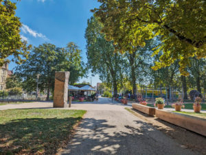 Stadtpark. Asphaltierter Weg im Schatten von Bäumen. Links ein Café mit großem Außenbereich. Rechts 2 Bänke aus zugesägten Holzstämmen. Rechts hinten ein Spielplatz.