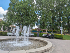 Im Vordergrund ein runder Springbrunnen. Um den Brunnen mehrere Sitzbänke. Im Hintergrund sehr große Bäume und in deren Schatten ein Spielplatz.