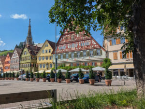 Malerischer Marktplatz mit historischen, farbenfrohen Fachwerkhäusern und einem gotischen Kirchturm im Hintergrund. Straßencafé mit Sonnenschirmen im Vordergrund, eingerahmt von Bäumen.