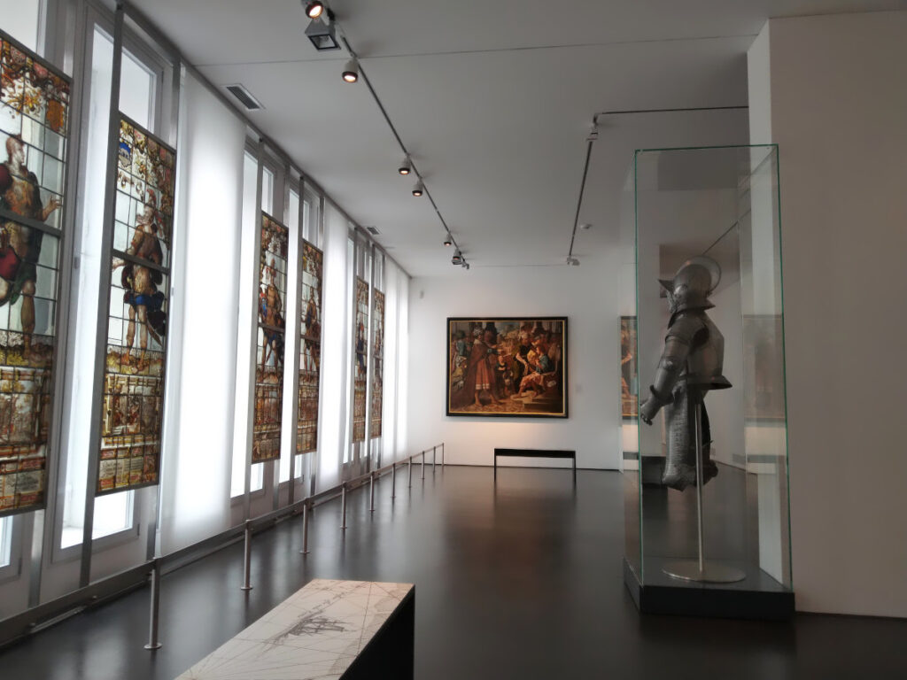 Gang mit Buntglasfenstern auf der linken Seite, die historische Personen zeigen. Rechts eine Vitrine mit einer Rüstung, geradeaus ein Gemälde.