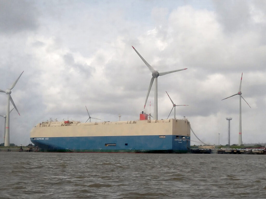 Großes Schiff ohne Fenster, unten blau und weiß, am Kai vertäut. Im Hintergrund sind mehrere Windräder zu sehen.