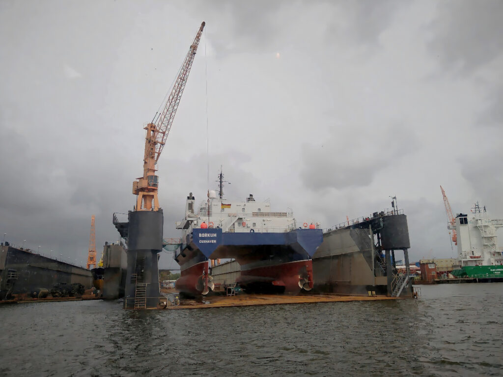 Schiff im schwimmenden Trockendock. Die Unterseite des Schiffs ist sichtbar. Besonders ist, dass es relativ flach ist und an beiden Seiten jeweils ein tiefen Rumpf mit einer Antriebsschraube ausgestattet ist.