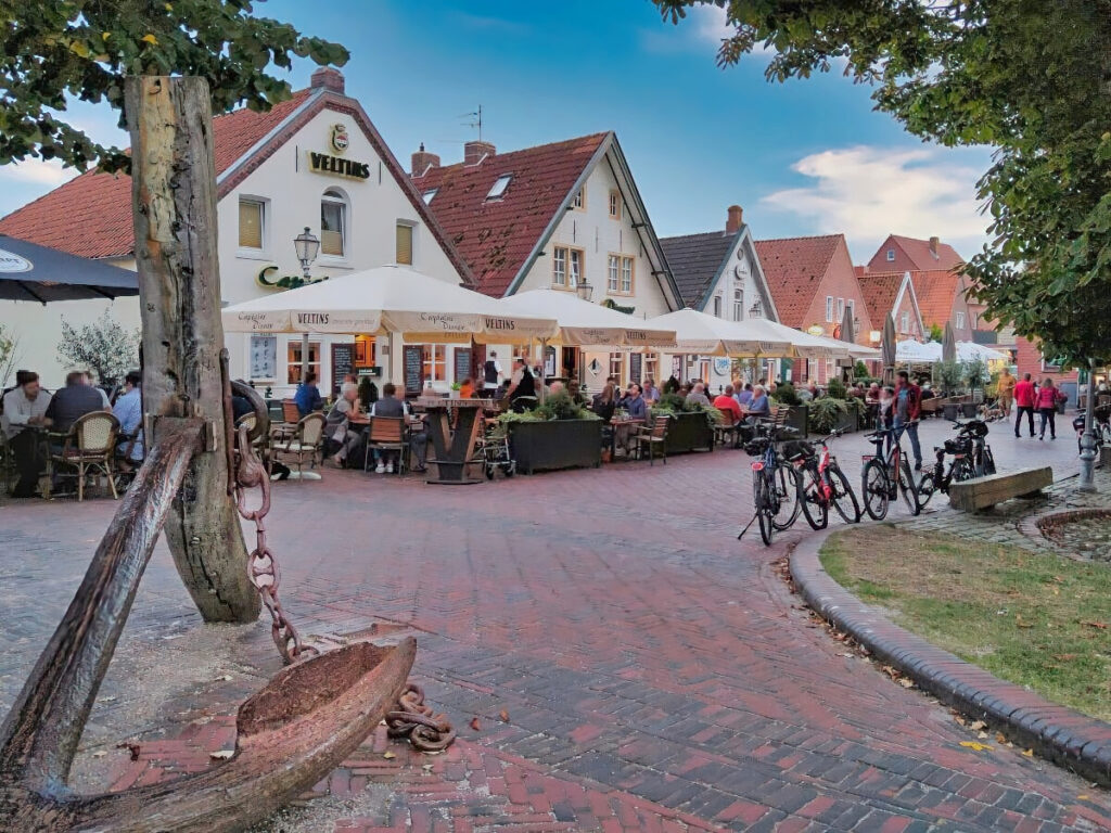 Fußgängerzone mit rotem und schwarzem Backsteinpflaster, gesäumt von zahlreichen Restaurants mit Außenbereichen. Links im Bild steht ein großer Anker als Skulptur auf der Straße.