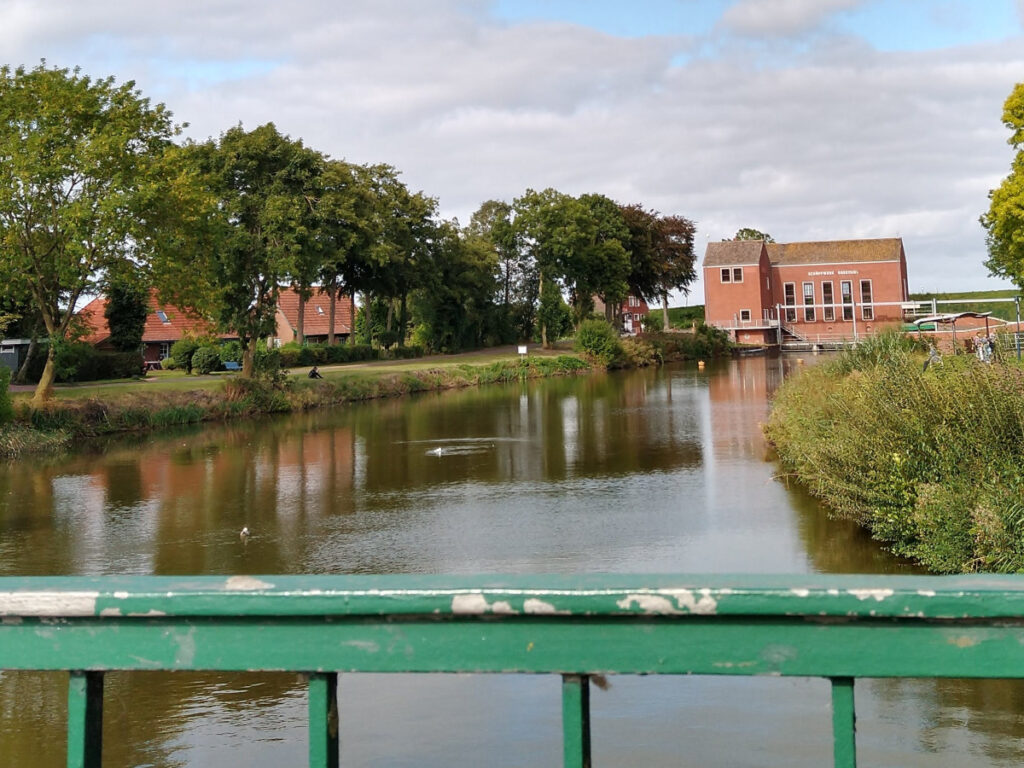 Blick von einer Brücke entlang eines breiten, mit Wasser gefüllten Kanals. Am Ende des Kanals steht ein Gebäude aus rotem Sandstein, hinter dem sich eine Grasdüne erstreckt.