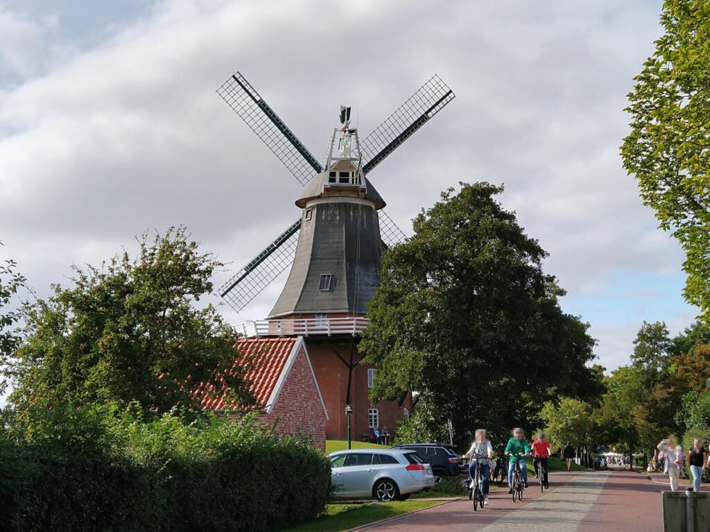 Links eine Windmühle mit rotem Backsteinbau im unteren Bereich, darüber eine graugrüne Holz-Etage und eine grüne Kappe mit Flügeln. Rechts eine Straße, auf der drei Radfahrer fahren.