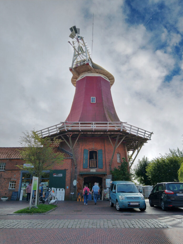 Längliches Backsteingebäude mit einem Aufbau einer roten Windmühle ohne Flügel auf der rechten Seite. Im flachen Gebäudeteil links befindet sich ein Laden. Vor der Mühle parken Autos.