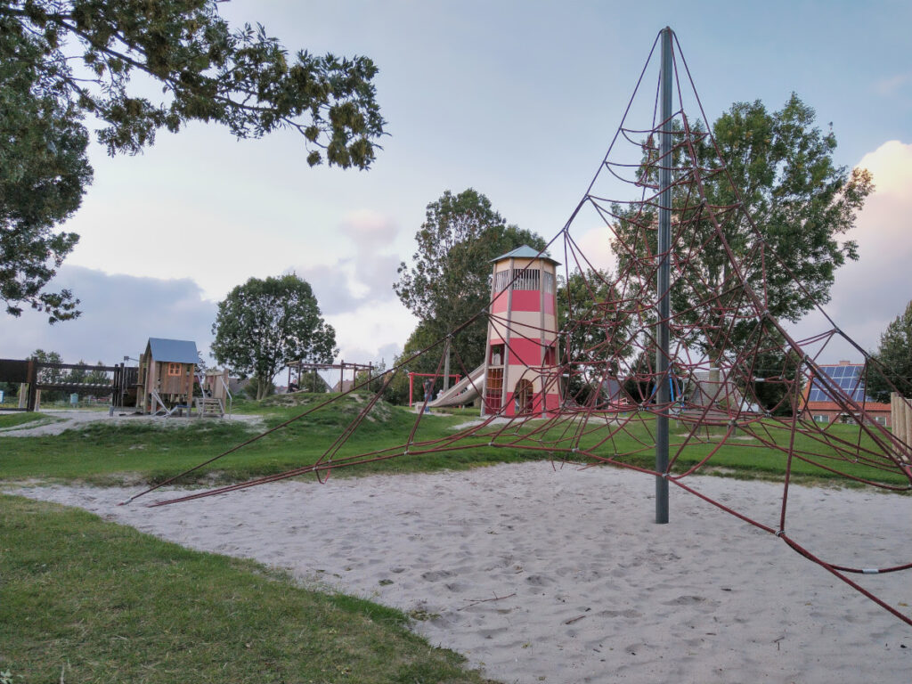 Spielplatz mit Klettergerüst in einer Sandgrube, Spielgebäude in Leuchtturmform mit Rutsche, Holzhäuschen und umliegender Wiese.