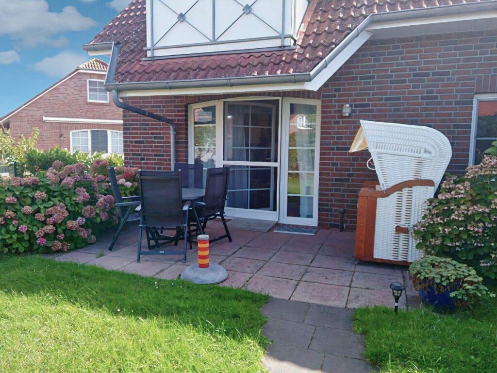 Blick vom Garten auf eine Terrasse mit einem runden Tisch und vier Stühlen auf der linken Seite, sowie einem Strandkorb auf der rechten Seite.