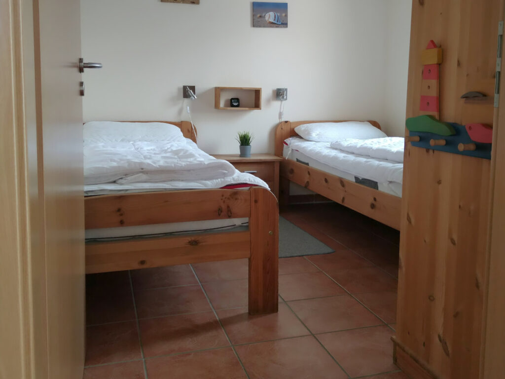 Blick ins Schlafzimmer. Links und rechts je ein Bett, dazwischen ein Gang. Rechts direkt nach der Tür ein Kleiderschrank, der den Durchgang verengt.