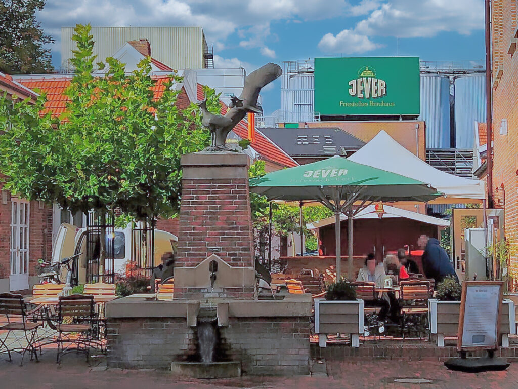 Blick aus einem Hinterhof mit rotem Backsteinbrunnen auf Silos mit einem großen Schild „Jever Friesisches Brauhaus“. Hinter dem Brunnen ein kleiner Außenbereich eines Restaurants, in dem vier Personen sitzen.