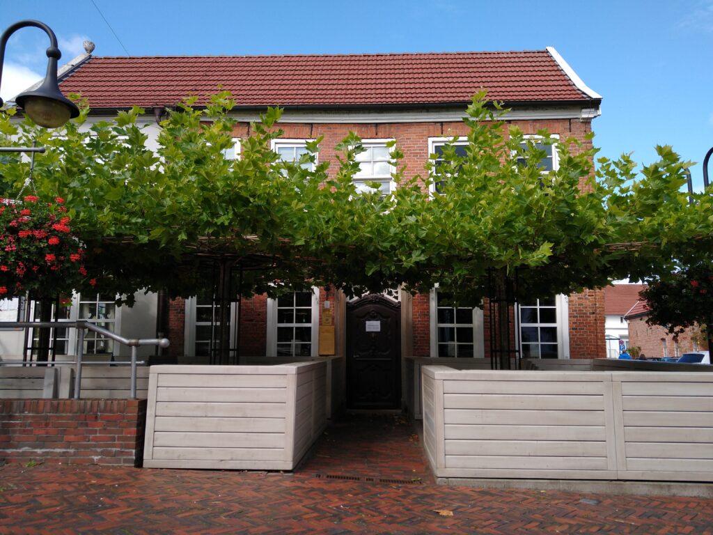 Zweigeschossiges Gebäude aus rotem Backstein. Davor ein Biergarten mit Bäumen, die eine dichte Laubkrone bilden.