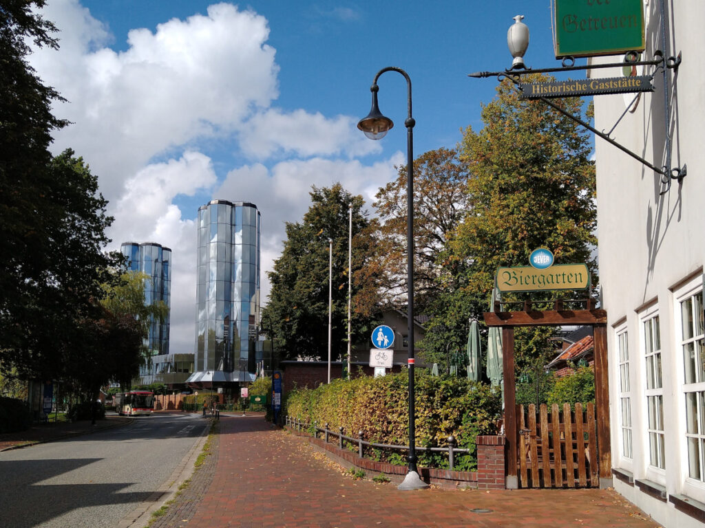 Links eine Straße, die zu zwei hohen, den Himmel reflektierenden Bürotürmen führt. Rechts ein Fußweg, an dessen rechter Seite sich ein Biergarten befindet.