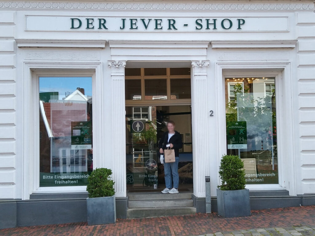 Weißes Gebäude mit Dekosäule am Eingang. Der Eingang ist über drei Stufen erreichbar und trägt den Schriftzug „Der Jever-Shop“. Rechts und links davon befinden sich Schaufenster.