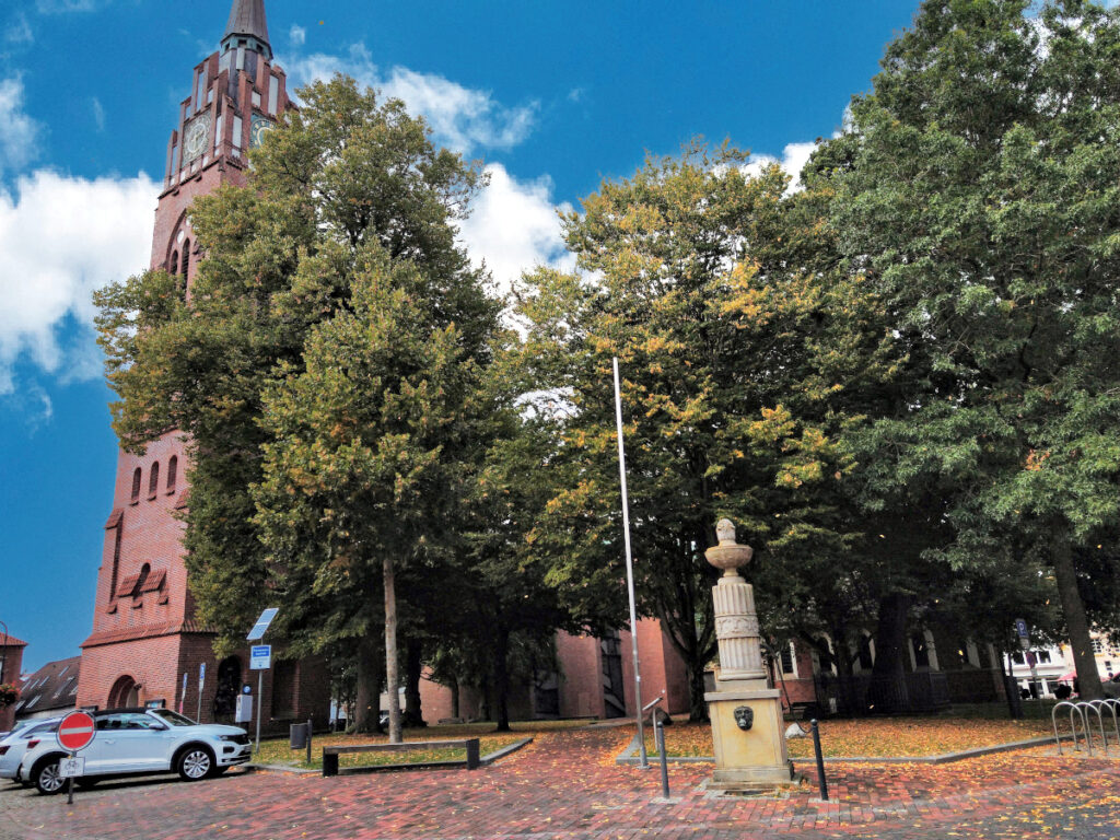 Kirche aus rotem Backstein, teilweise verdeckt durch drei große Bäume. Sichtbar ist hauptsächlich der Kirchturm auf der linken Seite.