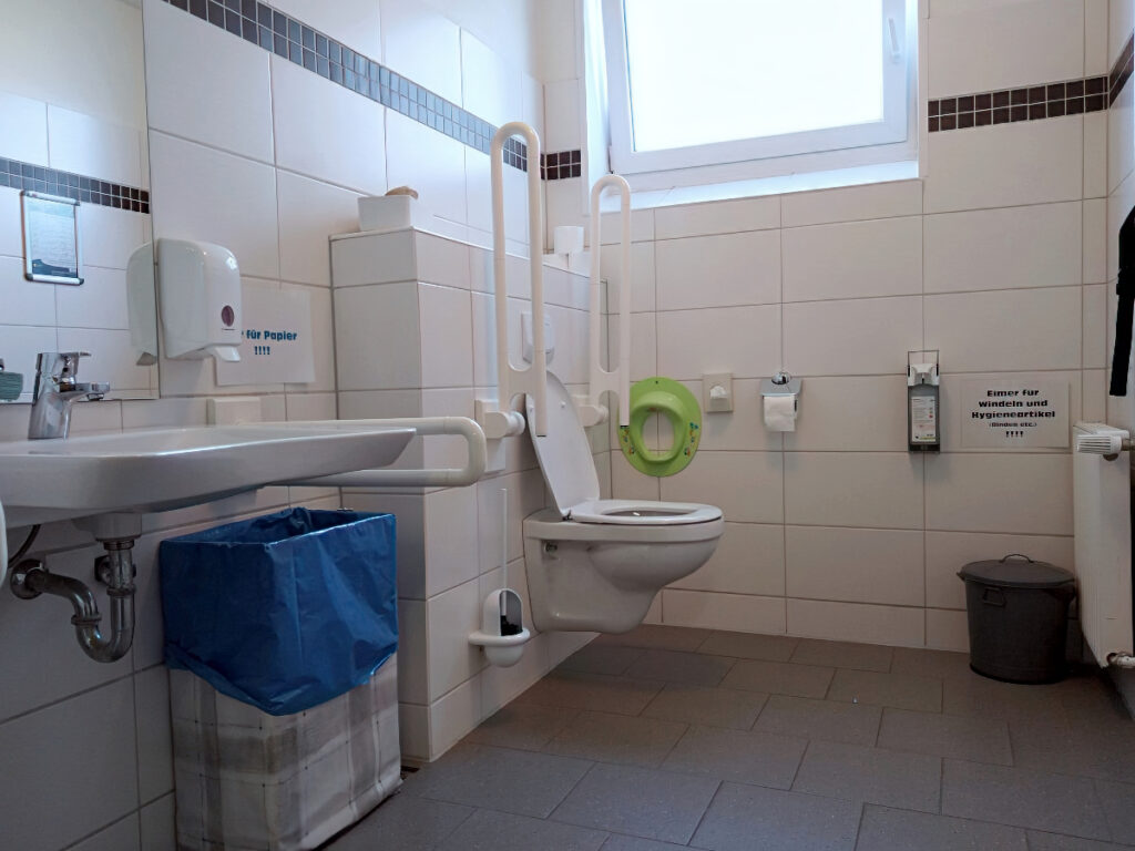 Barrierefreie Toilette mit einem Waschbecken vorne links und einem WC hinten links, ausgestattet mit beidseitigen Klappgriffen. Das WC befindet sich am hinteren Ende des Raums.