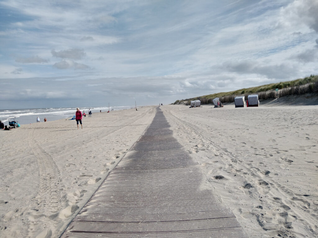 Weg aus Querplanken mit feinem Sandstrand auf beiden Seite. Der Weg führt weit den Sandstrand entlang. Links das Meer. Weniger Menschen sind am Strand.