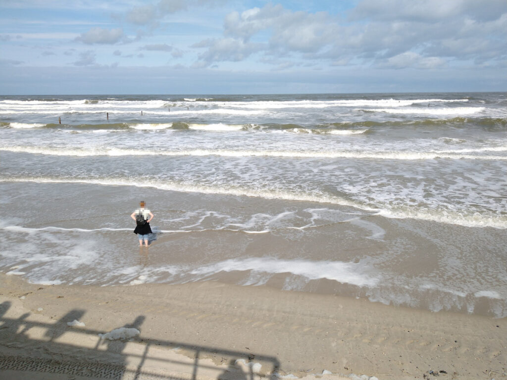 Blick von erhöht auf den Sandstrand: Eine Frau steht am Ufer und lässt ihre Füße vom Meer umspülen. In der Ferne ist der Horizont zu sehen.