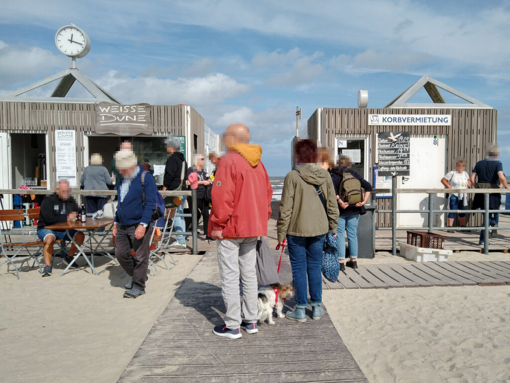 Plankenweg, der über den Sandstrand zu einer Plattform mit einem Kioskgebäude und einem Gebäude für die Strandkorbvermietung führt. Viele Menschen gehen den Weg entlang. Rechts führt ein Abzweig zum Strand.