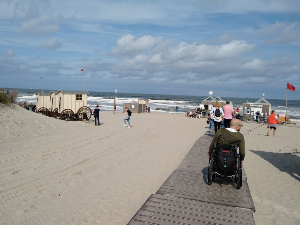 1,5 Meter breiter Plankenweg über den Sandstrand. Links Umkleidekabinen, der Weg führt zu einer Plattform mit zwei Gebäuden. Mehrere Personen sind über den Strand verteilt