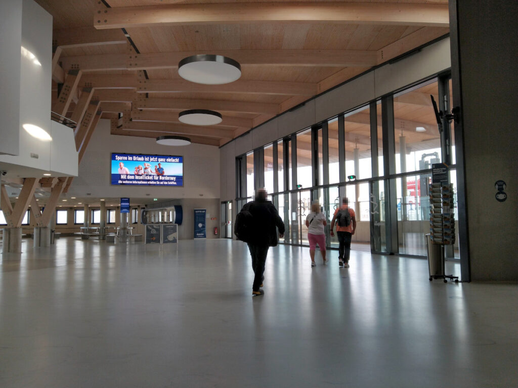 Große, weitläufige Halle. Im Hintergrund ein Wartebereich mit Sitzbänken, rechts mehrere elektrische Glastüren mit nur eingeschränktem Kontrast.