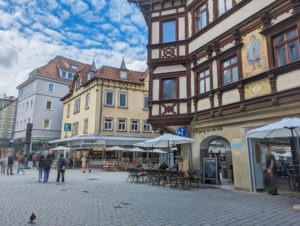 Gepflasterte Einkaufsstraße. Rechts ein Restaurant in einem historischen Fachwerkhaus, auf dessen Fassade sich ein Gemälde von einem Mann in einem mittelalterlichen Gewand befindet.