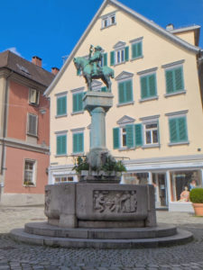 Runder Brunnen mit 2 Stufen. Auf dem Brunnenrand ein Relief mit Menschen in einem Wirtshaus. In der Mitte des Brunnen ragt eine Säule empor auf der ein Reiter steht, der in ein Horn bläst.