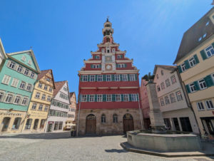 Rathausplatz mit dem Alten Rathaus. Im Vordergrund ein Brunnen mit Adlerfigur, umgeben von Fachwerkhäusern unter blauem Himmel. Platz mit Kopfsteinpflaster.