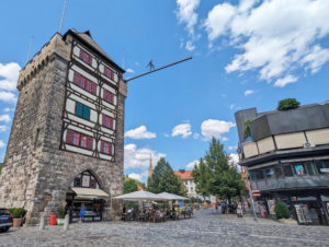 Links mittelalterlicher Turm aus groben Steinen. Auf einer Seite sind mittig 4 Etagen nicht mit Mauerwerk, sondern mit Fachwerk gemacht. Mittig Fußgängerzone. Rechts modernes Gebäude.