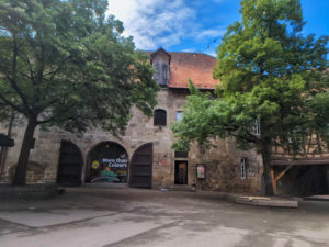 Schattiger Hof mit 2 großen Bäumen. Im Hintergrund ein aus groben Steinen gemauertes Gebäude mit einem großem Fenster und einer geöffneten Tür mit 3 Stufen.