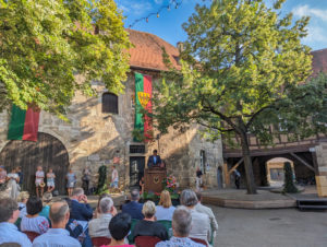 Innenhof mit historischen Gebäude und 2 Bäumen. Im Hof sitzen mehrere Personen und hören einem Mann an einem Rednerpult zu.